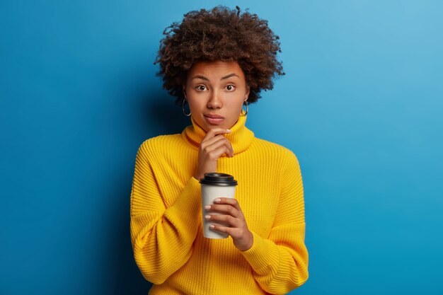 Afroamerikanerin gekleidet in gelbem Pullover, trinkt Kaffee zum Mitnehmen, hält Tasse vom Café