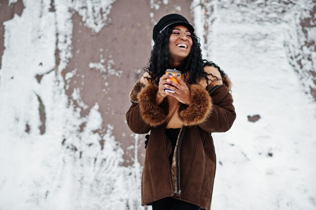 Afroamerikanerin in Schaffellmantel und Mütze posierte am Wintertag vor schneebedecktem Hintergrund mit einer Tasse Kaffee