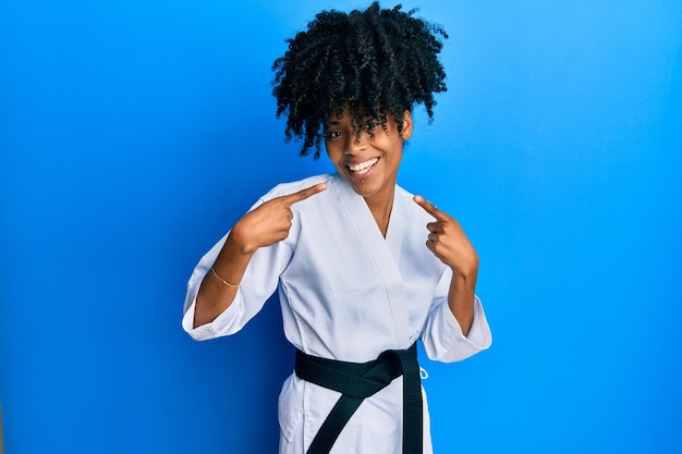 Afroamerikanerin mit afro-haar trägt karate-kimono und schwarzen gürtel und lächelt fröhlich, zeigt und zeigt mit fingern, zähnen und mund. zahngesundheitskonzept.