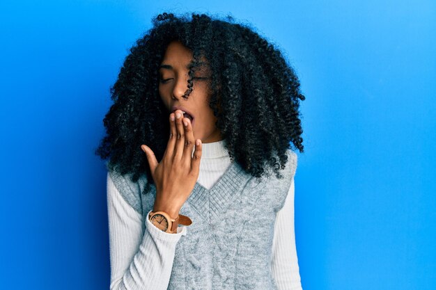 Afroamerikanerin mit Afro-Haaren, die einen lässigen Winterpullover trägt, gelangweilt gähnend und müde, den Mund mit der Hand bedeckend. Unruhe und Schläfrigkeit.