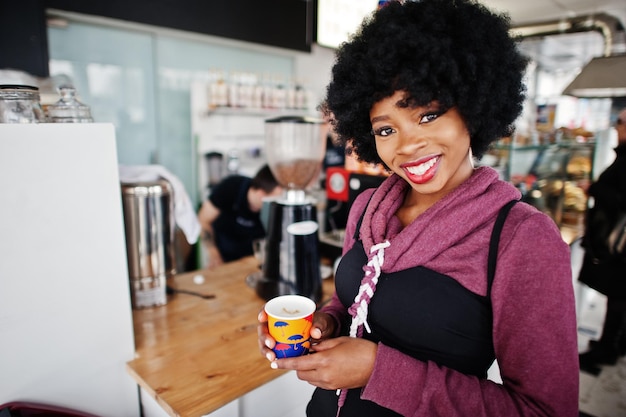 Afroamerikanerin mit lockigem Haar trägt einen Pullover, der im Café drinnen mit einer Tasse Tee oder Kaffee posiert