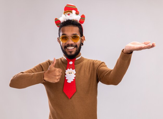 Afroamerikanermann in braunem Pullover und Weihnachtsmannrand auf Kopf mit lustiger roter Krawatte, die Daumen oben zeigt, die mit Arm lächelnd fröhlich stehend über weißem Hintergrund präsentieren