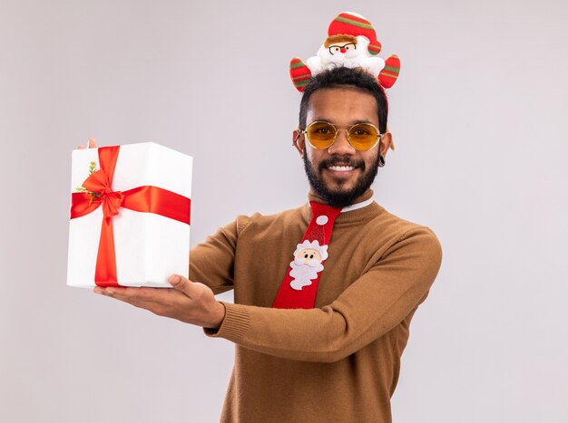 Afroamerikanermann in braunem Pullover und Weihnachtsmannrand auf Kopf mit lustiger roter Krawatte, die ein Geschenk hält, das Kamera mit Lächeln auf Gesicht steht, das über weißem Hintergrund steht