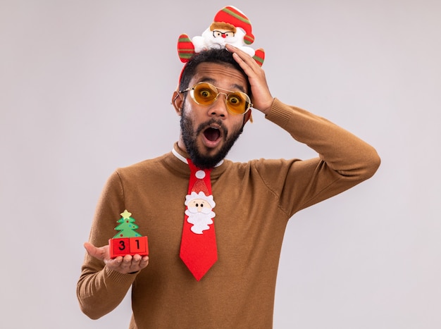 Afroamerikanermann in braunem Pullover und Weihnachtsmannrand auf Kopf mit lustiger roter Krawatte, die Spielzeugwürfel mit Neujahrsdatum betrachtet, das Kamera betrachtet und überrascht über weißem Hintergrund steht