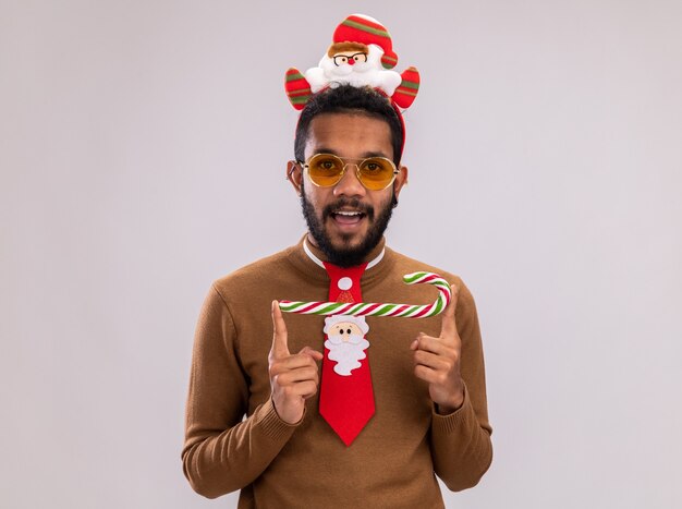 Afroamerikanermann in braunem Pullover und Weihnachtsmannrand auf Kopf mit lustiger roter Krawatte, die Zuckerstange hält, die Kamera betrachtet, die fröhlich über weißem Hintergrund steht