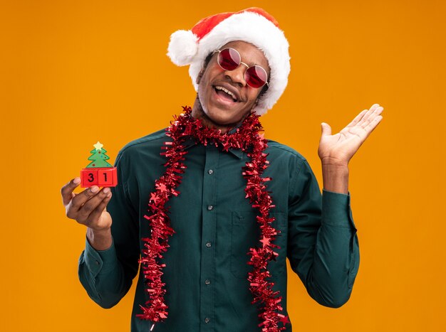 Afroamerikanermann in der Weihnachtsmannmütze mit Girlande, die Sonnenbrille hält, die Spielzeugwürfel mit Neujahrsdatum glücklich und fröhlich mit erhöhtem Arm hält, der über orange Wand steht