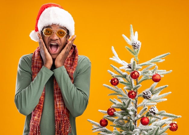 Afroamerikanermann in der Weihnachtsmannmütze und im Schal um den Hals, der Kamera glücklich und aufgeregt steht, steht neben einem Weihnachtsbaum über orange Hintergrund