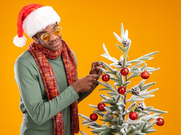 Afroamerikanermann in der Weihnachtsmannmütze und im Schal um Hals hängenden Weihnachtskugeln auf einem Weihnachtsbaum glücklich und positiv stehend über orange Wand