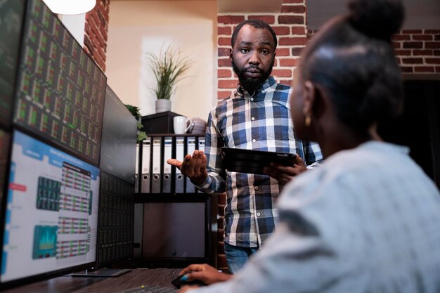 Afroamerikanische Finanzinvestoren in Büroarbeitsplätzen diskutieren über geringe Gewinnchancen. Forex-Börsenhändler sitzt an einer Workstation mit mehreren Monitoren, während er mit einem Kollegen spricht.