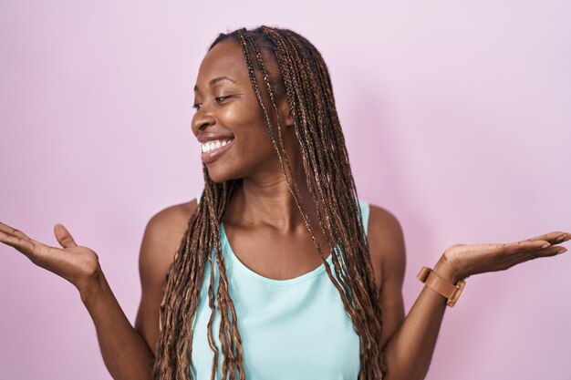 Afroamerikanische Frau steht lächelnd vor rosa Hintergrund und zeigt mit beiden Händen offene Handflächen, um Vergleiche und Balance zu präsentieren und zu werben