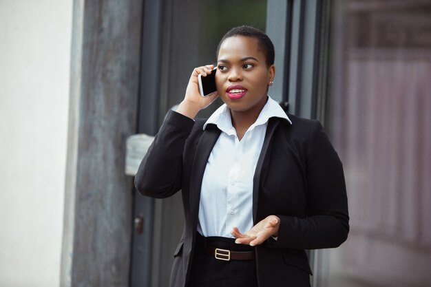 Afroamerikanische Geschäftsfrau in Bürokleidung lächelnd sieht selbstbewusst am Telefon sprechen