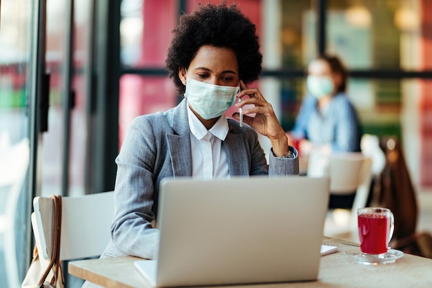 Afroamerikanische Geschäftsfrau trägt Schutzmaske, während sie mit dem Handy kommuniziert und in einem Café am Laptop arbeitet