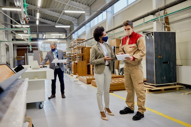 Afroamerikanische Geschäftsfrau und männlicher Arbeiter tragen Gesichtsmasken, während sie Berichte in der Holzfabrik prüfen