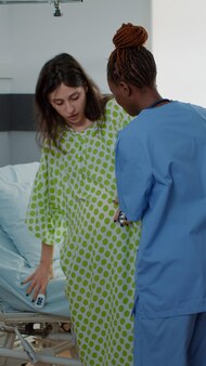 Afroamerikanische krankenschwester, die schwangere frau unterstützt, lag im krankenbett. patient erwartet baby in der entbindungsklinik mit gesundheitsgeräten und medizinischem personal. junge kaukasische mutter