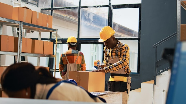Afroamerikanische Männer verpacken Produkte in Kartons und planen mit Lagerwerkzeugen den Einzelhandelsversand im Lagerraum. Männliche Angestellte, die in der Produktlogistik und im Warenversand arbeiten. Handaufnahme.
