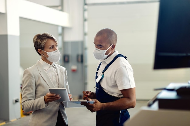 Afroamerikanische Mechanikerin und Geschäftsfrau, die Gesichtsmasken tragen, während sie das Touchpad in einer Reparaturwerkstatt verwenden