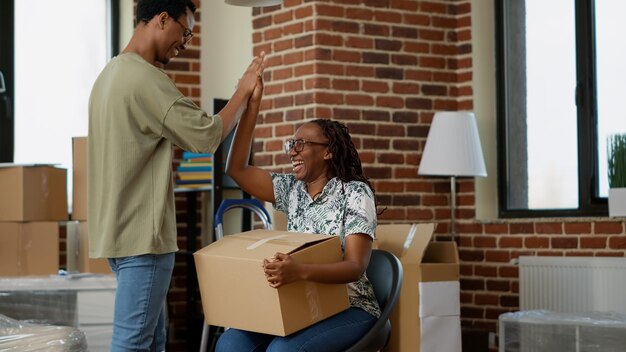 Afroamerikanische Partner haben Spaß mit ausgepackten Möbeln in einer neuen Haushaltswohnung. Umzüge genießen, zusammen in eine Mietwohnung ziehen und mit Deko rumalbern. Handschuss.