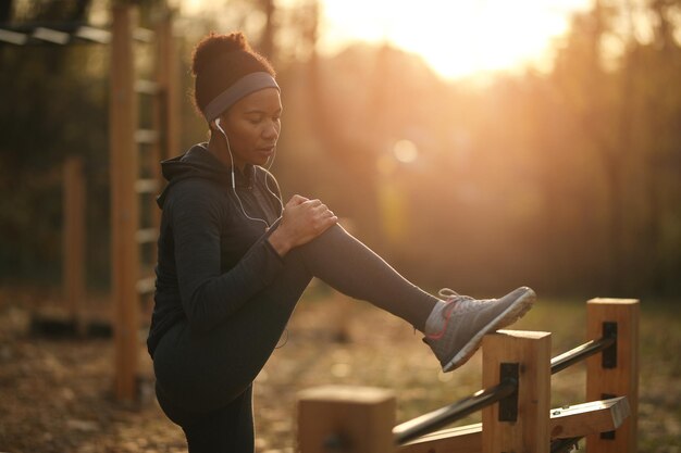 Afroamerikanische Sportlerin, die ihr Knie vor Schmerzen hält, während sie bei Sonnenuntergang im Park trainiert