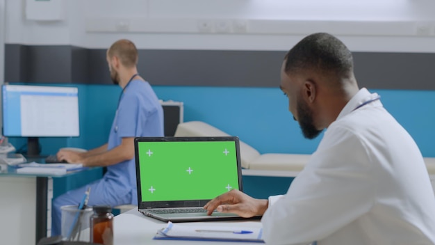 Afroamerikanischer arzt arzt, der sich einen greenscreen-chroma-key-laptop-computer mit isolierter anzeige ansieht, der medizinisches fachwissen im krankenhausbüro eingibt. medizinkonzept