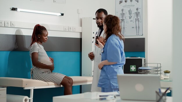 Afroamerikanischer Arzt, der einer schwangeren Patientin bei einem Kontrollbesuch verschreibungspflichtige Pillen gibt. Männlicher Arzt hilft Frau mit Schwangerschaftsbauch und zeigt Flasche mit Medikamentenbehandlung.
