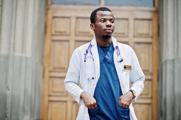 Afroamerikanischer Arzt männlich im Laborkittel mit Stethoskop im Freien gegen Kliniktür