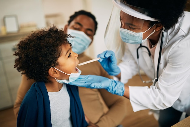 Afroamerikanischer Arzt mit Gesichtsmaske, der bei einem Hausbesuch den Hals des Jungen untersucht