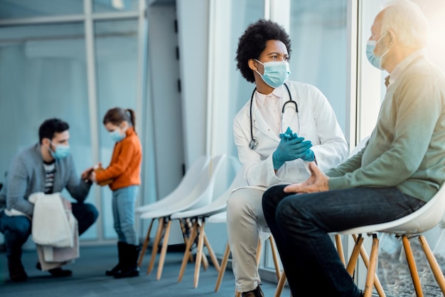 Afroamerikanischer Arzt und älterer Mann kommunizieren im Wartezimmer des Krankenhauses, während sie Gesichtsmasken tragen