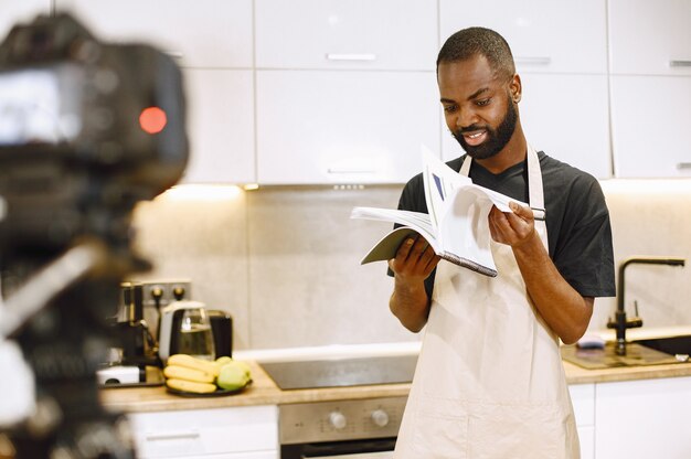 Afroamerikanischer bärtiger Mann, der ein Kochbuch lächelt und liest. Blogger-Aufnahmevideo zum Kochen von Vlog in der Küche zu Hause. Mann trägt eine Schürze.