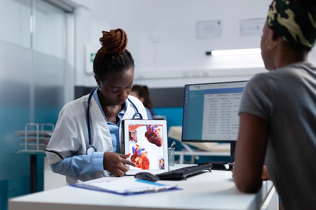 Afroamerikanischer Fachkardiologe, der einem kranken Patienten das Fachwissen über die Herzradiographie erklärt und die medikamentöse Behandlung während des klinischen Termins bespricht. Arztfrau, die im Krankenhausbüro arbeitet