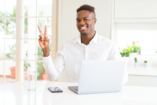 afroamerikanischer Geschäftsmann, der mit einem Laptop arbeitet, lächelt mit einem glücklichen Gesicht und zwinkert in die Kamera, während er das Siegeszeichen Nummer zwei macht