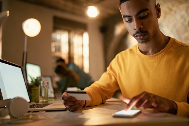 Afroamerikanischer Geschäftsmann mit Kreditkarte und Handy, während er sein Online-Bankkonto während der Nachtarbeit im Büro überprüft