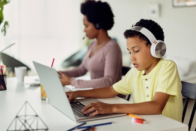 Afroamerikanischer Junge mit Laptop, während seine Mutter im Hintergrund arbeitet