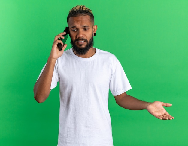 Afroamerikanischer junger Mann im weißen T-Shirt, der beim Telefonieren verwirrt aussieht