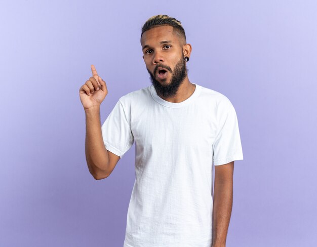 Afroamerikanischer junger Mann im weißen T-Shirt, der die Kamera anschaut, überrascht mit dem Zeigefinger, der eine neue großartige Idee hat, die auf blauem Hintergrund steht