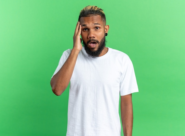 Afroamerikanischer junger Mann im weißen T-Shirt, der die Kamera erstaunt und überrascht auf grünem Hintergrund betrachtet