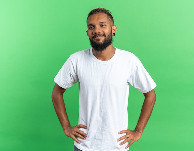 Afroamerikanischer junger Mann im weißen T-Shirt, der die Kamera mit einem Lächeln im Gesicht mit Armen an der Hüfte über grünem Hintergrund anschaut