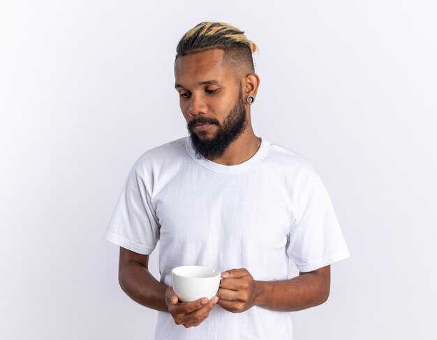 Afroamerikanischer junger Mann im weißen T-Shirt, der eine Tasse heißen Tee hält und mit traurigem Ausdruck beiseite schaut