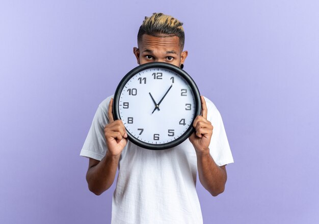 Afroamerikanischer junger Mann im weißen T-Shirt, der eine Wanduhr vor seinem Gesicht hält, besorgt