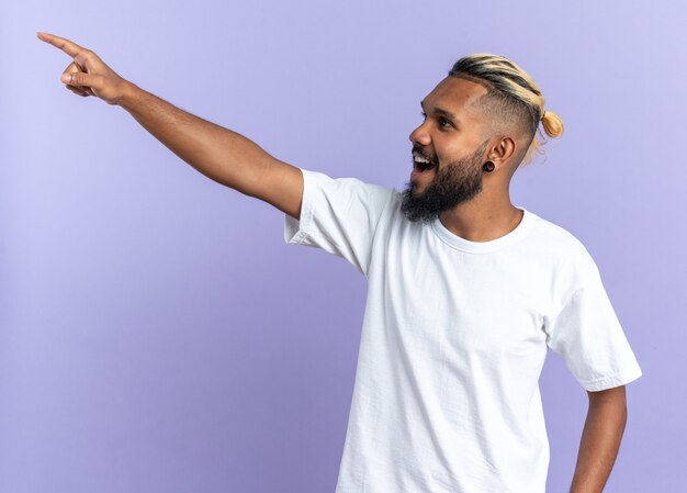 Afroamerikanischer junger Mann im weißen T-Shirt, der glücklich und fröhlich beiseite schaut und mit dem Zeigefinger auf etwas zeigt, das auf blauem Hintergrund steht