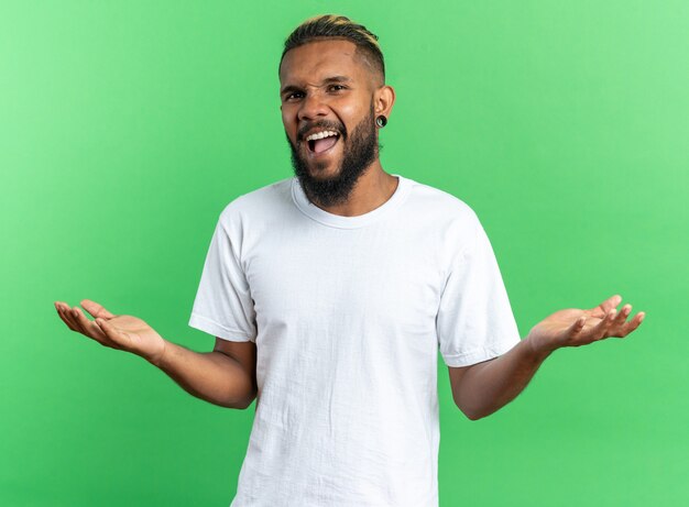 Afroamerikanischer junger Mann im weißen T-Shirt, der mit erhobenen Armen schreit, wütend und unzufrieden über grünem Hintergrund steht