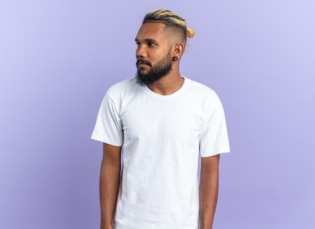 Afroamerikanischer junger Mann im weißen T-Shirt, der mit ernstem Gesicht auf blauem Hintergrund beiseite schaut
