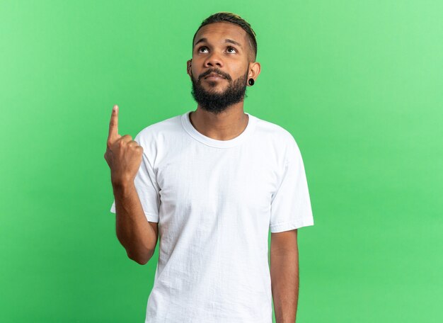 Afroamerikanischer junger Mann im weißen T-Shirt, der mit ernstem Gesicht aufschaut und mit dem Zeigefinger auf etwas zeigt, das über grünem Hintergrund steht