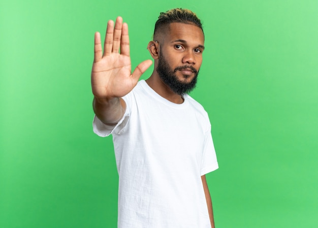 Afroamerikanischer junger mann im weißen t-shirt, der mit ernstem gesicht in die kamera schaut und eine stopp-geste mit der hand über grünem hintergrund macht