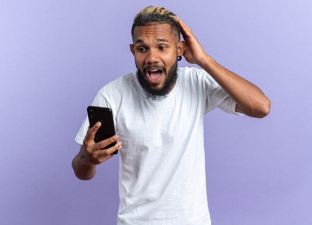 Afroamerikanischer junger Mann im weißen T-Shirt, der Smartphone hält und es mit der Hand auf dem Kopf schockiert, die über blauem Hintergrund steht