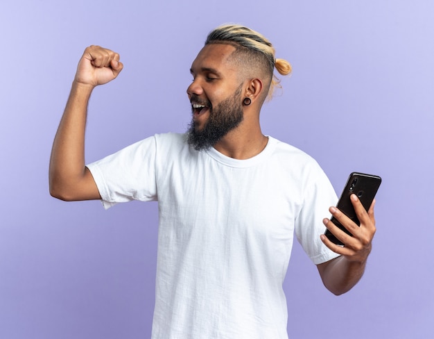 Afroamerikanischer junger Mann im weißen T-Shirt, der Smartphone mit geballter Faust glücklich und aufgeregt schreit und sich über seinen Erfolg freut