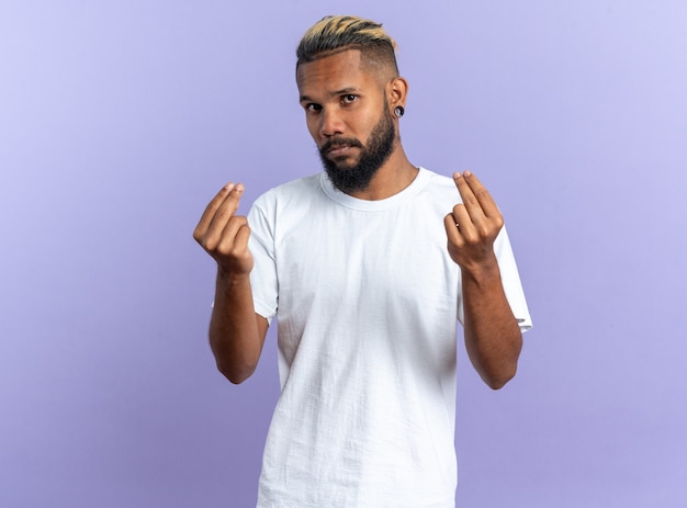 Afroamerikanischer junger Mann im weißen T-Shirt mit Blick auf die Kamera, die Geldgeste macht und die Finger auf blauem Hintergrund reibt