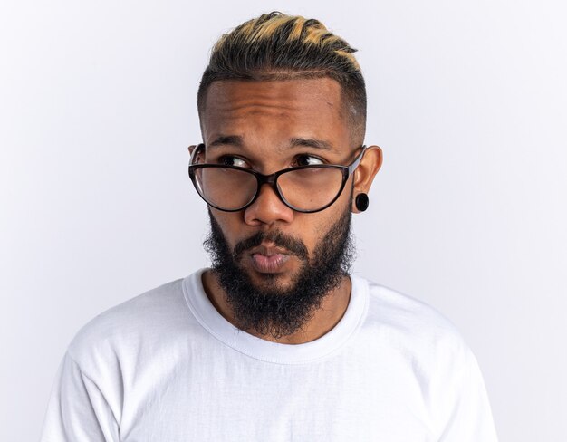 Afroamerikanischer junger Mann im weißen T-Shirt mit Brille, der verwirrt über Weiß steht