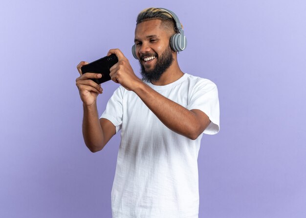 Afroamerikanischer junger Mann im weißen T-Shirt mit Kopfhörern, die Spiele mit Smartphone spielen, glücklich und aufgeregt