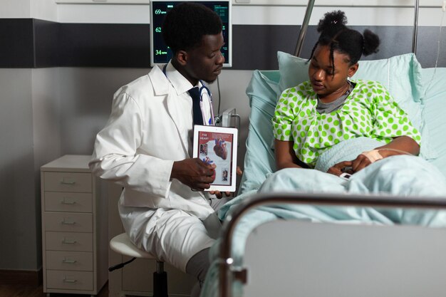 Afroamerikanischer Kardiologe, der einem kranken Patienten eine Tablette mit Herzröntgen hält. Therapeut bespricht die medizinische Behandlung während der Konsultation auf der Krankenstation. Kardiogramm auf dem Bildschirm