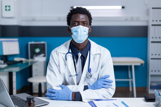 Afroamerikanischer Krankenhausarzt, der wegen einer Coronavirus-Pandemie am Schreibtisch sitzt und eine Gesichtsmaske trägt. Klinikarzt sitzt am Schreibtisch und trägt Virenschutzmaske